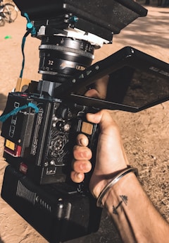 A hand is holding a professional film camera with visible lens and monitor. The camera's design includes various buttons, dials, and a large lens marked with the brand 'ARRI'. A bracelet and tattoo are visible on the person's wrist. The background is outdoors, with a dirt ground and hints of other equipment.