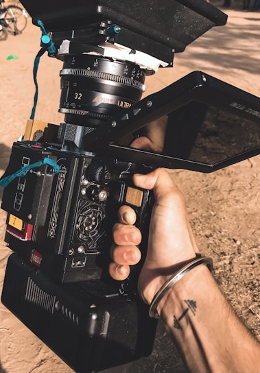 A hand is holding a professional film camera with visible lens and monitor. The camera's design includes various buttons, dials, and a large lens marked with the brand 'ARRI'. A bracelet and tattoo are visible on the person's wrist. The background is outdoors, with a dirt ground and hints of other equipment.