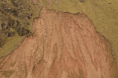A steep slope with exposed reddish-brown soil and scattered rocks, bordered by a contrasting green, grassy area. The slope appears eroded, likely caused by natural processes.