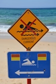 A warning sign on a beach indicates a dangerous current with a red line crossing out a swimming figure on a yellow triangular sign. Below it, a blue rectangular sign with swimming icon and flags suggests safe swimming areas to the left.