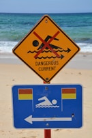 A warning sign on a beach indicates a dangerous current with a red line crossing out a swimming figure on a yellow triangular sign. Below it, a blue rectangular sign with swimming icon and flags suggests safe swimming areas to the left.