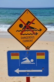 A warning sign on a beach indicates a dangerous current with a red line crossing out a swimming figure on a yellow triangular sign. Below it, a blue rectangular sign with swimming icon and flags suggests safe swimming areas to the left.