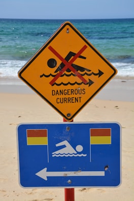 A warning sign on a beach indicates a dangerous current with a red line crossing out a swimming figure on a yellow triangular sign. Below it, a blue rectangular sign with swimming icon and flags suggests safe swimming areas to the left.