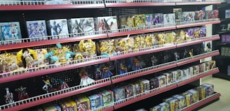Shelves filled with various toy figurines and boxed sets, organized neatly. The figurines, mostly golden and shiny, depict various characters in dynamic poses. The boxed sets include colorful and detailed packaging featuring imagery of the characters. The overall arrangement is orderly, displaying a collection of collectibles.