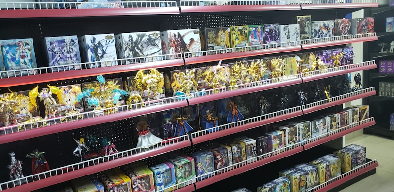 Shelves filled with various toy figurines and boxed sets, organized neatly. The figurines, mostly golden and shiny, depict various characters in dynamic poses. The boxed sets include colorful and detailed packaging featuring imagery of the characters. The overall arrangement is orderly, displaying a collection of collectibles.