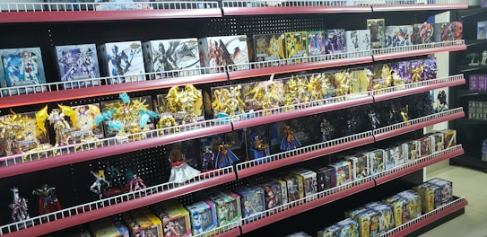 Shelves filled with various toy figurines and boxed sets, organized neatly. The figurines, mostly golden and shiny, depict various characters in dynamic poses. The boxed sets include colorful and detailed packaging featuring imagery of the characters. The overall arrangement is orderly, displaying a collection of collectibles.