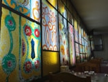 A series of stained glass panels with intricate geometric and floral patterns are illuminated from behind. The designs feature bold colors including red, yellow, green, and blue, with each panel showcasing unique artistic elements. Wooden chairs and tables with glasses are seen in the foreground, suggesting a dining setting.
