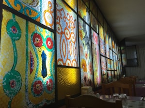 A series of stained glass panels with intricate geometric and floral patterns are illuminated from behind. The designs feature bold colors including red, yellow, green, and blue, with each panel showcasing unique artistic elements. Wooden chairs and tables with glasses are seen in the foreground, suggesting a dining setting.