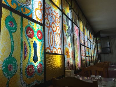 A series of stained glass panels with intricate geometric and floral patterns are illuminated from behind. The designs feature bold colors including red, yellow, green, and blue, with each panel showcasing unique artistic elements. Wooden chairs and tables with glasses are seen in the foreground, suggesting a dining setting.