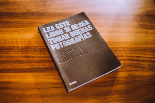 A book with a dark cover is placed on a wooden surface. The cover text reads 'Lea este libro si desea tomar buenas fotografías' by Henry Carroll. The background consists of a rich, warm wood grain texture.