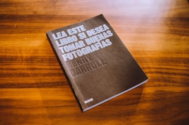 A book with a dark cover is placed on a wooden surface. The cover text reads 'Lea este libro si desea tomar buenas fotografías' by Henry Carroll. The background consists of a rich, warm wood grain texture.