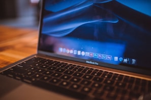 A close-up view of a MacBook Pro laptop with a blurred wooden table in the background. The screen displays a macOS desktop with several Adobe Creative Cloud applications visible in the dock, including Illustrator, Photoshop, and Premiere Pro. The lighting gives the scene a warm and cozy atmosphere.
