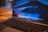 A close-up view of a MacBook Pro laptop with a blurred wooden table in the background. The screen displays a macOS desktop with several Adobe Creative Cloud applications visible in the dock, including Illustrator, Photoshop, and Premiere Pro. The lighting gives the scene a warm and cozy atmosphere.
