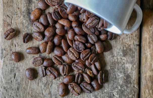 A collection of roasted coffee beans spills out of a white ceramic mug onto a rustic wooden surface. The beans are rich brown with a glossy sheen, highlighting their texture and detailing.