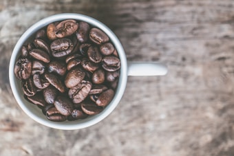 A white cup filled with dark brown coffee beans is placed on a textured wooden surface. The image is taken from a top-down perspective, highlighting the rich details of the coffee beans and their glossy textures.
