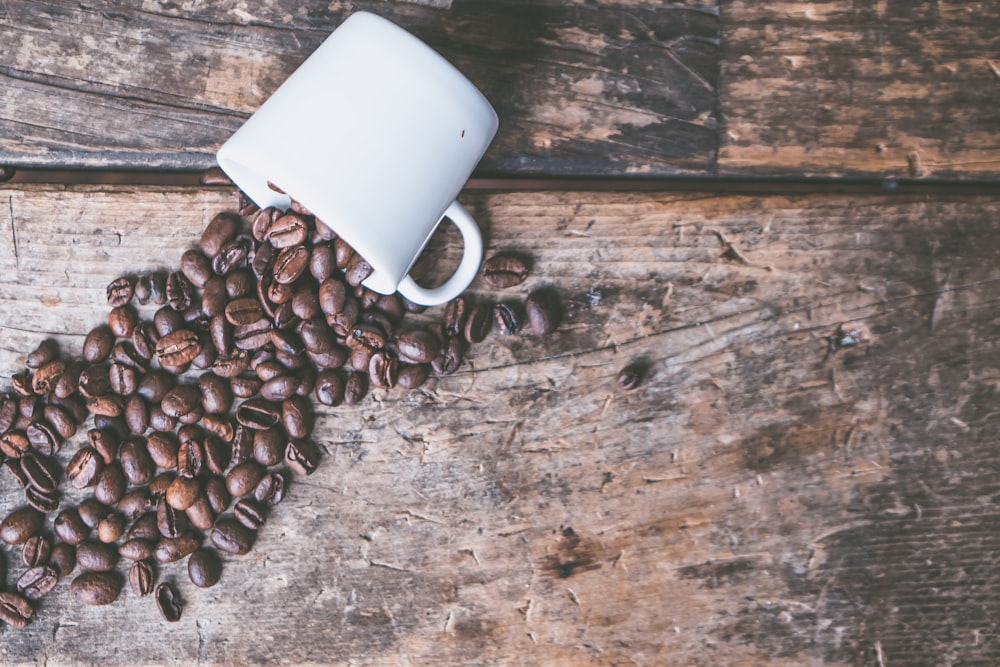 coffee beans spill on brown wooden surface from white mug