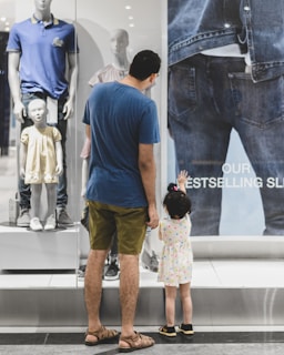 A man and a small child stand in front of a store display window. The man is wearing a blue shirt and khaki shorts, while the child is in a polka-dot dress reaching up towards the glass. The display features mannequins dressed in casual clothing and a large poster advertising jeans.