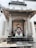 A colorful statue of a traditional Chinese deity is seated within an ornate stone shrine. The deity is depicted in vibrant robes, holding a symbolic object. The shrine is adorned with stone carvings and Chinese characters on pillars. The ground in front of the statue has incense sticks.