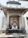 A colorful statue of a traditional Chinese deity is seated within an ornate stone shrine. The deity is depicted in vibrant robes, holding a symbolic object. The shrine is adorned with stone carvings and Chinese characters on pillars. The ground in front of the statue has incense sticks.