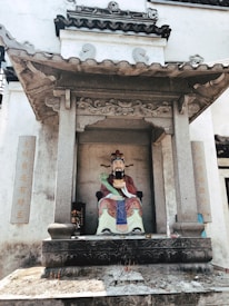 A colorful statue of a traditional Chinese deity is seated within an ornate stone shrine. The deity is depicted in vibrant robes, holding a symbolic object. The shrine is adorned with stone carvings and Chinese characters on pillars. The ground in front of the statue has incense sticks.
