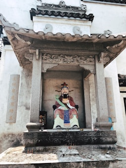 A colorful statue of a traditional Chinese deity is seated within an ornate stone shrine. The deity is depicted in vibrant robes, holding a symbolic object. The shrine is adorned with stone carvings and Chinese characters on pillars. The ground in front of the statue has incense sticks.