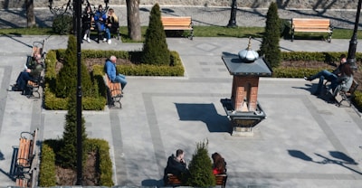A small public park area featuring several benches and landscaped greenery surrounding a central fountain structure. People are seated on the benches, some engaged in conversation, others relaxing alone. The park is well-maintained with paved walkways and neatly trimmed bushes.