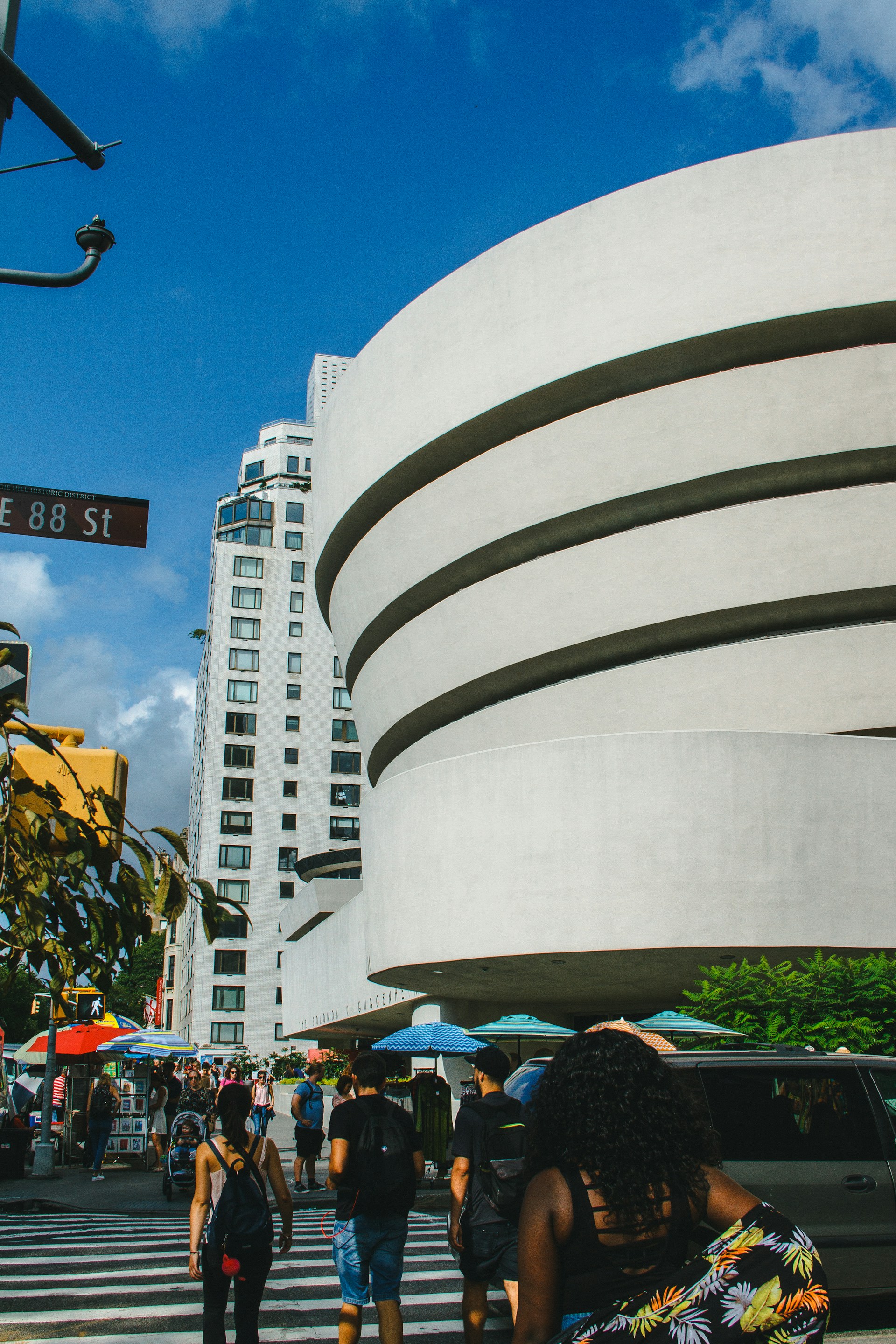 A bustling urban scene features a modern, curved architectural building with distinct horizontal lines. People are walking along the crosswalk in varied attire, suggesting an active and lively environment. Tall city buildings dominate the background against a vibrant blue sky.