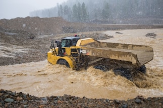 A construction vehicle, specifically a dump truck, is navigating through a flooded area with muddy water. The landscape is rough and mountainous, with a backdrop of dense trees shrouded in mist. Heavy rainfall is visible, creating a harsh and challenging environment.