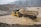 A construction vehicle, specifically a dump truck, is navigating through a flooded area with muddy water. The landscape is rough and mountainous, with a backdrop of dense trees shrouded in mist. Heavy rainfall is visible, creating a harsh and challenging environment.