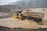 A construction vehicle, specifically a dump truck, is navigating through a flooded area with muddy water. The landscape is rough and mountainous, with a backdrop of dense trees shrouded in mist. Heavy rainfall is visible, creating a harsh and challenging environment.