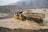 A construction vehicle, specifically a dump truck, is navigating through a flooded area with muddy water. The landscape is rough and mountainous, with a backdrop of dense trees shrouded in mist. Heavy rainfall is visible, creating a harsh and challenging environment.