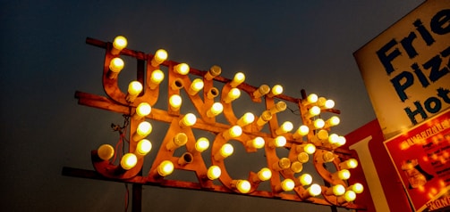 A brightly lit vintage sign with numerous round bulbs arranged in letters, set against a dark sky background. To the right, partially visible signage for food items including pizza and hotdogs.