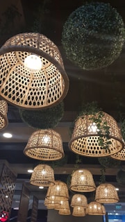 Rattan-style hanging lamps with intricate weaving patterns illuminate the space. Large, spherical decor made of artificial greenery is interspersed among the lights. The warm glow from the lamps creates a cozy and inviting atmosphere.
