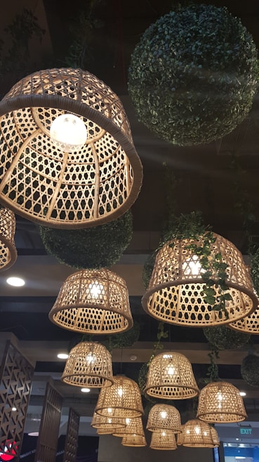 Rattan-style hanging lamps with intricate weaving patterns illuminate the space. Large, spherical decor made of artificial greenery is interspersed among the lights. The warm glow from the lamps creates a cozy and inviting atmosphere.