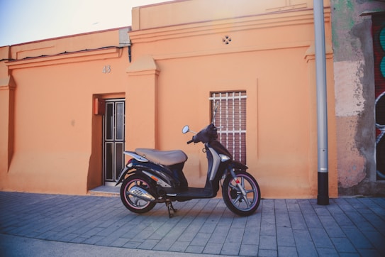 A black motor scooter is parked on a paved sidewalk in front of an orange building. The building has a door marked with the number 43 and a window with metal bars. There is a streetlamp nearby and part of a mural is visible on the adjacent wall.