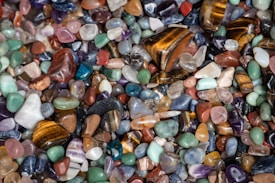 A collection of polished gemstones in various shapes and sizes, exhibiting a wide range of colors including greens, blues, reds, browns, and purples. The stones are closely packed together, creating a visually rich mosaic of textures and hues.