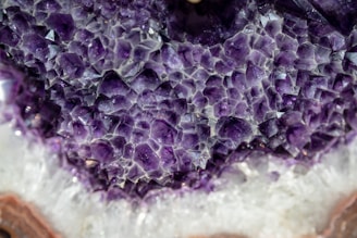 A close-up view of a geode with a dazzling array of purple amethyst crystals. The crystals are jagged and multifaceted, glistening as the light catches their surfaces. Surrounding the purple sections, there are white and brown layers that add contrast and depth.