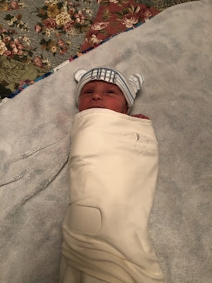 A newborn baby is swaddled in a soft cream-colored blanket, lying on a bed with a floral-patterned quilt. The baby is wearing a striped hat with small ears and the word 'CUTIE' embroidered on it, adding a playful touch.