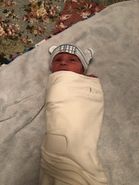 A newborn baby is swaddled in a soft cream-colored blanket, lying on a bed with a floral-patterned quilt. The baby is wearing a striped hat with small ears and the word 'CUTIE' embroidered on it, adding a playful touch.