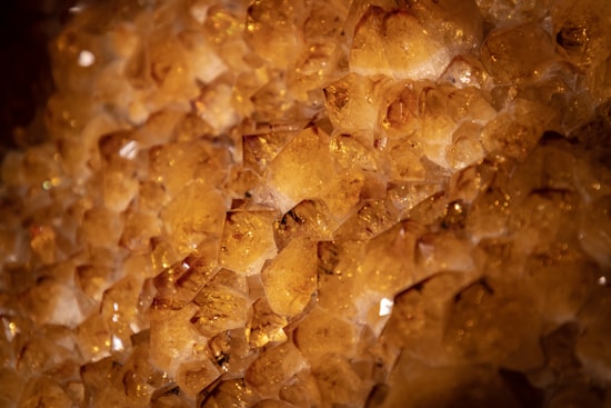 A close-up view of a cluster of citrine crystals, displaying their sharp angular surfaces and rich honey-gold coloration. The light reflects off the facets, highlighting their translucent quality and creating an intricate pattern of shapes and textures.