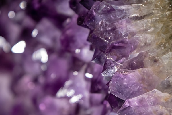 Close-up view of an array of purple amethyst crystals displaying a variety of hues from deep violet to light lavender, with a hint of white reflections. The crystals have a rough, jagged texture and the light interacts with their surfaces to create a shimmering effect.