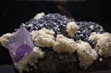 A cluster of minerals is displayed, featuring large crystalline structures. The prominent mineral is a purple cubic crystal, likely a fluorite, surrounded by a mix of dark black, metallic-looking rocks and white, chalky formations.