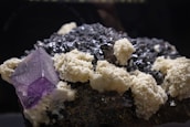 A cluster of minerals is displayed, featuring large crystalline structures. The prominent mineral is a purple cubic crystal, likely a fluorite, surrounded by a mix of dark black, metallic-looking rocks and white, chalky formations.
