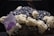 A cluster of minerals is displayed, featuring large crystalline structures. The prominent mineral is a purple cubic crystal, likely a fluorite, surrounded by a mix of dark black, metallic-looking rocks and white, chalky formations.