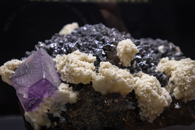 A cluster of minerals is displayed, featuring large crystalline structures. The prominent mineral is a purple cubic crystal, likely a fluorite, surrounded by a mix of dark black, metallic-looking rocks and white, chalky formations.