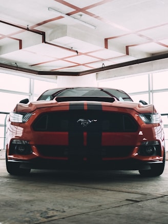 A muscular red sports car with black racing stripes is parked indoors, likely in a garage or showroom. The car has a distinctive front end with sharp headlights and a prominent logo on the grille. The surrounding environment is industrial, featuring exposed pipes and a concrete ceiling with visible trusses.