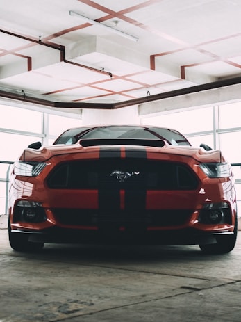A muscular red sports car with black racing stripes is parked indoors, likely in a garage or showroom. The car has a distinctive front end with sharp headlights and a prominent logo on the grille. The surrounding environment is industrial, featuring exposed pipes and a concrete ceiling with visible trusses.