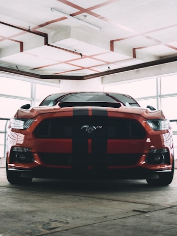A muscular red sports car with black racing stripes is parked indoors, likely in a garage or showroom. The car has a distinctive front end with sharp headlights and a prominent logo on the grille. The surrounding environment is industrial, featuring exposed pipes and a concrete ceiling with visible trusses.