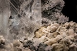 A close-up of a crystalline structure showcasing intricate, clear crystal formations intertwined with rough, rocky surfaces. Smaller crystal clusters with a sparkling appearance are visible, surrounded by a mix of smoother and jagged textures.