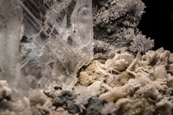 A close-up of a crystalline structure showcasing intricate, clear crystal formations intertwined with rough, rocky surfaces. Smaller crystal clusters with a sparkling appearance are visible, surrounded by a mix of smoother and jagged textures.